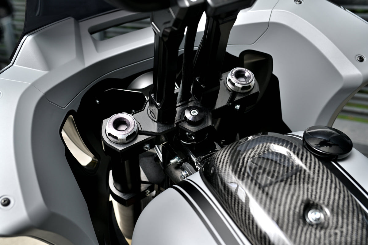 Close-up detail of a motorcycle's handlebar area, showing black handlebar mounts, silver bolts, a carbon fiber tank cover, and light gray body panels.