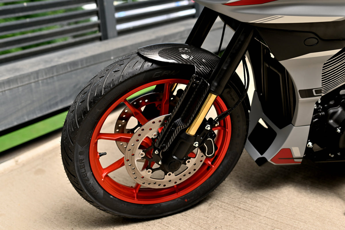 Close-up of a motorcycle's front wheel featuring a black tire, a carbon fiber front fender, a black fork with gold accents, and a bright orange-red wheel rim.
