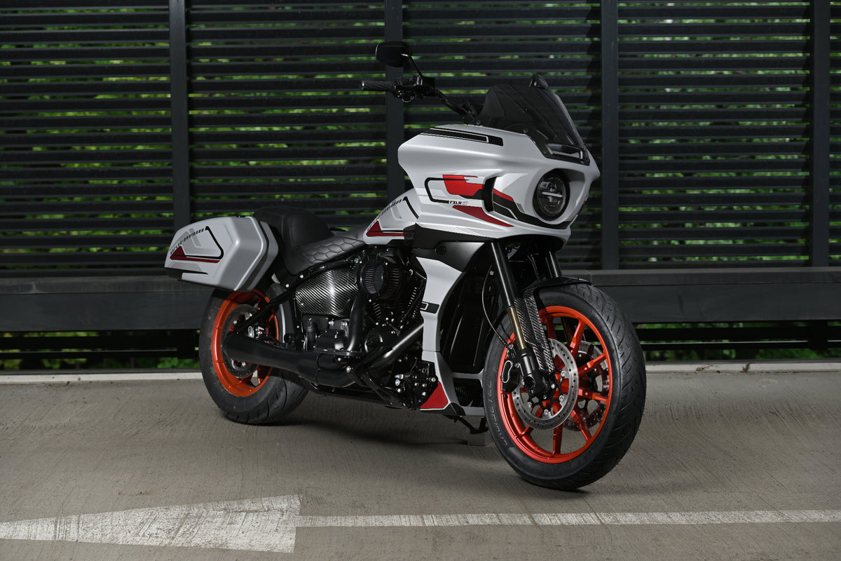A silver and black Harley Davidson FXLRST with vibrant orange-red wheel accents, parked on a concrete surface in front of a dark green slatted wall.