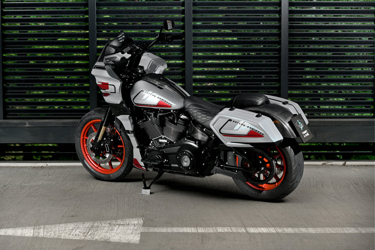 A Harley-Davidson Low Rider ST with a silver and black body, red wheels, and carbon fiber parts, parked on a concrete surface in front of a dark green slatted wall.