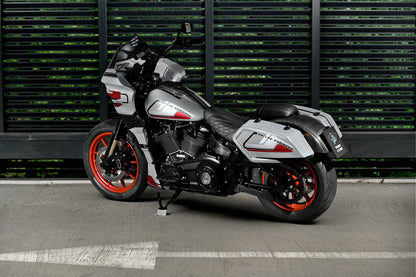 A Harley-Davidson Low Rider ST with a silver and black body, red wheels, and carbon fiber parts, parked on a concrete surface in front of a dark green slatted wall.
