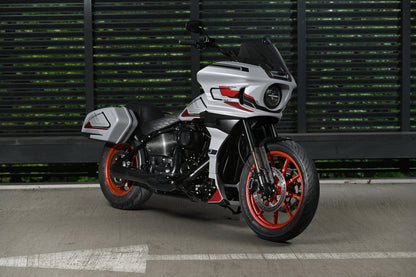A silver and black Harley Davidson FXLRST with vibrant orange-red wheel accents, parked on a concrete surface in front of a dark green slatted wall.